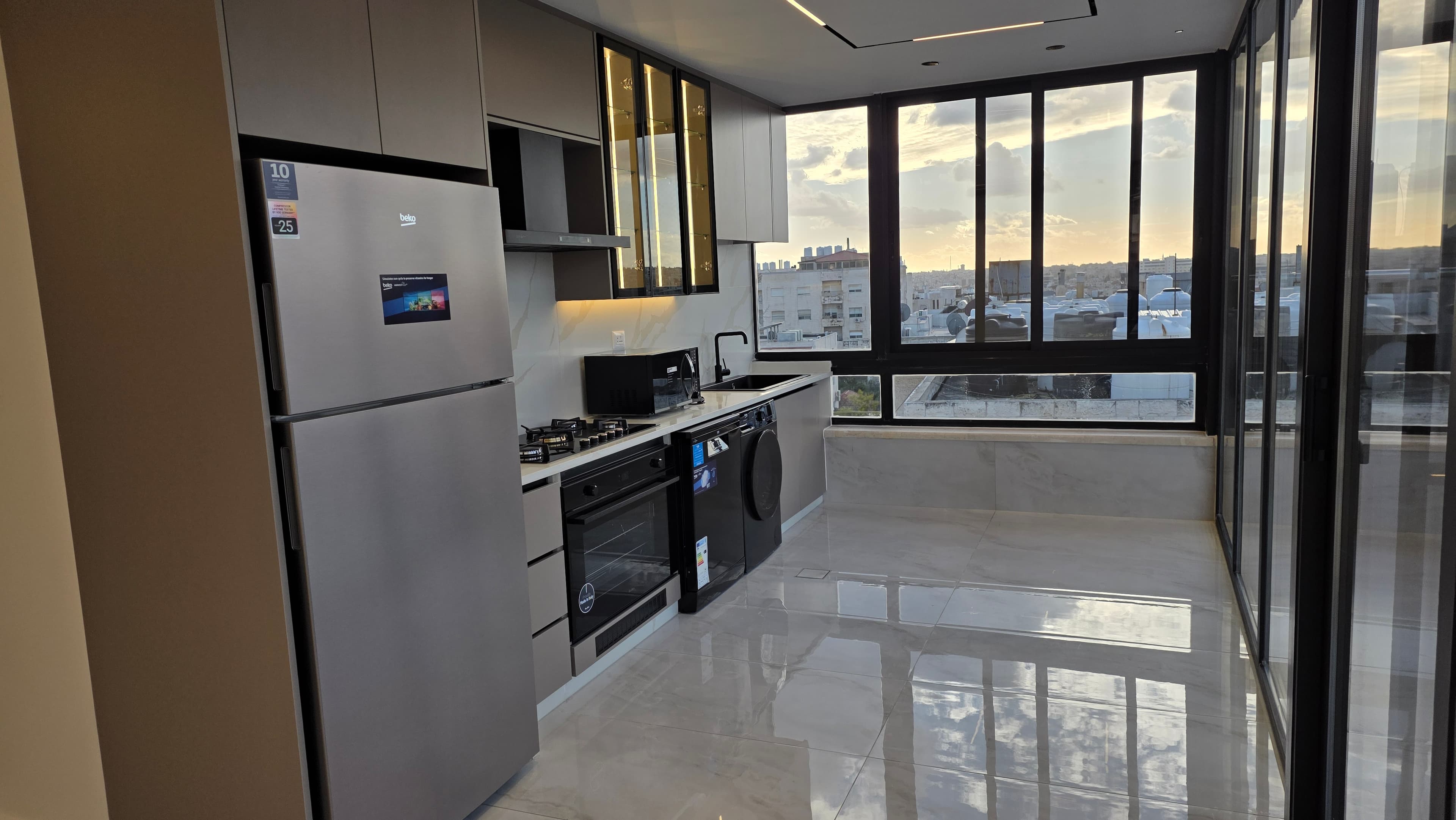 Galley-style kitchen at golden hour with Beko fridge, matte taupe cabinetry, smoked glass upper cabinets with LED interiors, Calacatta marble backsplash, corner windows with skyline views, terrace sliding doors